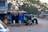 A small roadside stall with a blue and palm-leaf roof stands next to a deserted road. An auto-rickshaw with a heart symbol painted on its side is parked near the stall. The background features tall trees and a few distant vehicles.