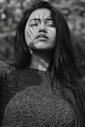 A black and white portrait of a person with long hair, eyes closed, and a serene expression. A shadow of a branch with leaves is cast over the face, adding an artistic element.