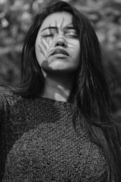 A black and white portrait of a person with long hair, eyes closed, and a serene expression. A shadow of a branch with leaves is cast over the face, adding an artistic element.