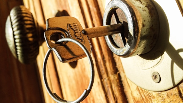A close-up of a metal key inserted into a slightly rusted lock on a wooden door. The surrounding area shows details of the lock and door texture, with sunlight casting shadows.