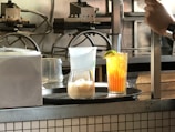 A kitchen or bar counter setup with two drinks displayed in the foreground. One is a light-colored drink with ice in a rounded glass, and the other is a vibrant orange beverage with a lime wedge garnish in a tall glass. Visible behind the drinks are some appliances and machinery, possibly for making waffles.