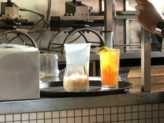 A kitchen or bar counter setup with two drinks displayed in the foreground. One is a light-colored drink with ice in a rounded glass, and the other is a vibrant orange beverage with a lime wedge garnish in a tall glass. Visible behind the drinks are some appliances and machinery, possibly for making waffles.