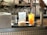 A kitchen or bar counter setup with two drinks displayed in the foreground. One is a light-colored drink with ice in a rounded glass, and the other is a vibrant orange beverage with a lime wedge garnish in a tall glass. Visible behind the drinks are some appliances and machinery, possibly for making waffles.