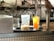 A kitchen or bar counter setup with two drinks displayed in the foreground. One is a light-colored drink with ice in a rounded glass, and the other is a vibrant orange beverage with a lime wedge garnish in a tall glass. Visible behind the drinks are some appliances and machinery, possibly for making waffles.