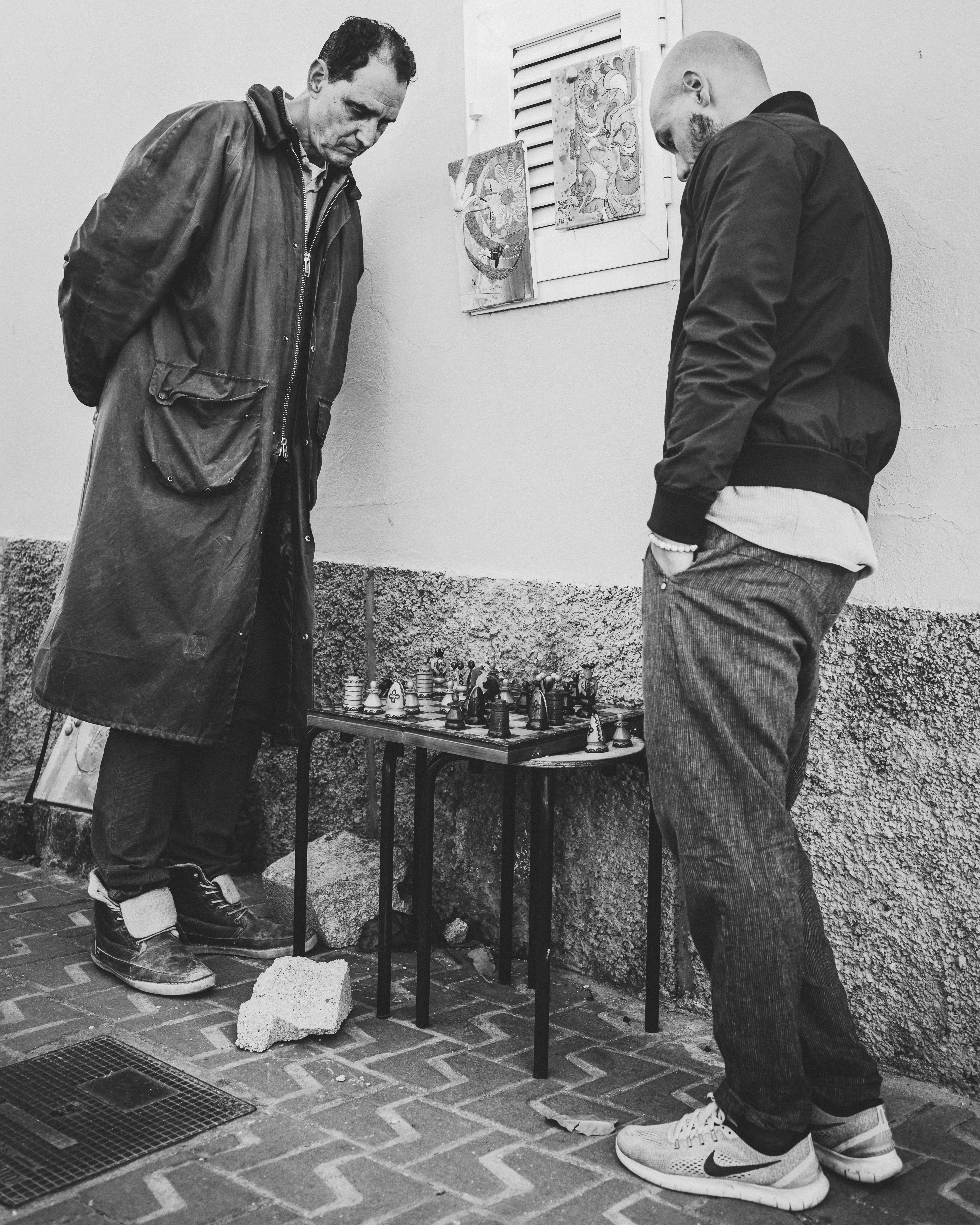 Two men stand intently over a small chessboard, which is placed on a folding table against a textured wall. Both are dressed in casual clothing; one wears a long coat and the other a jacket and jeans. The setting appears to be outdoors, as indicated by the brick pavement beneath them. A couple of artworks or posters are attached to the wall behind them.