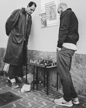 Two men stand intently over a small chessboard, which is placed on a folding table against a textured wall. Both are dressed in casual clothing; one wears a long coat and the other a jacket and jeans. The setting appears to be outdoors, as indicated by the brick pavement beneath them. A couple of artworks or posters are attached to the wall behind them.
