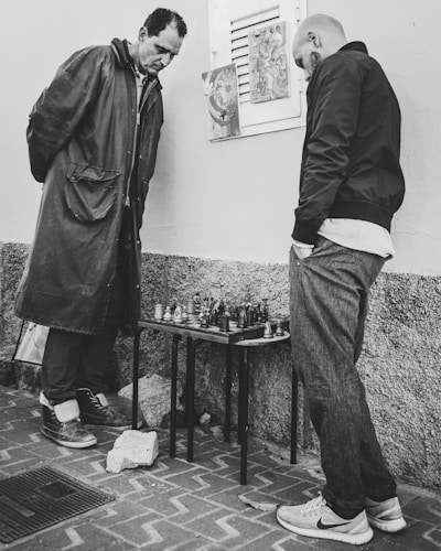Two men stand intently over a small chessboard, which is placed on a folding table against a textured wall. Both are dressed in casual clothing; one wears a long coat and the other a jacket and jeans. The setting appears to be outdoors, as indicated by the brick pavement beneath them. A couple of artworks or posters are attached to the wall behind them.