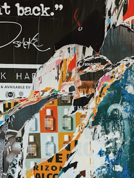 A collage of torn posters revealing layers of text, graphics, and fragmented images. The piece features a mix of colors, including blues, oranges, and whites. Some visible text includes phrases like 'SOLD OUT' and partial words, alongside graphics of small bottles.