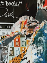 A collage of torn posters revealing layers of text, graphics, and fragmented images. The piece features a mix of colors, including blues, oranges, and whites. Some visible text includes phrases like 'SOLD OUT' and partial words, alongside graphics of small bottles.