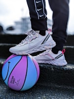 A pair of light gray sneakers with pink accents are resting on a colorful blue and purple basketball. The person wearing the sneakers is dressed in black pants with white stripes and a series of Nike logos. The scene appears to be on a set of steps or a similar textured surface.