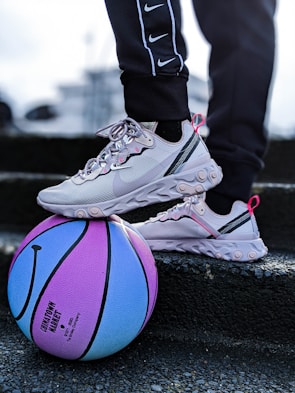 A pair of light gray sneakers with pink accents are resting on a colorful blue and purple basketball. The person wearing the sneakers is dressed in black pants with white stripes and a series of Nike logos. The scene appears to be on a set of steps or a similar textured surface.