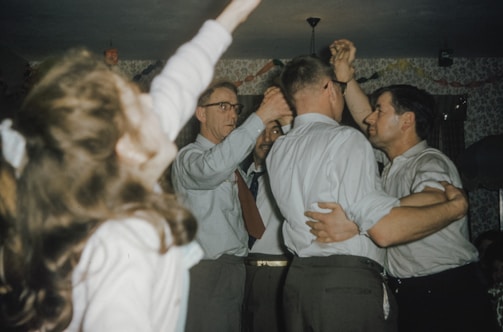A group of men is engaged in dancing or a festive activity, appearing formal in shirts and trousers. A young girl is in the foreground, also participating in the festivities. The setting suggests an indoor party with dim lighting and a warm, jovial atmosphere.