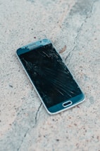 A smartphone with a cracked screen lies on a rough concrete surface. The cracks spread across most of the display, indicating significant damage.