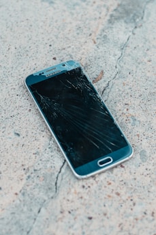A smartphone with a cracked screen lies on a rough concrete surface. The cracks spread across most of the display, indicating significant damage.