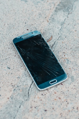 A smartphone with a cracked screen lies on a rough concrete surface. The cracks spread across most of the display, indicating significant damage.
