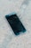 A smartphone with a cracked screen lies on a rough concrete surface. The cracks spread across most of the display, indicating significant damage.
