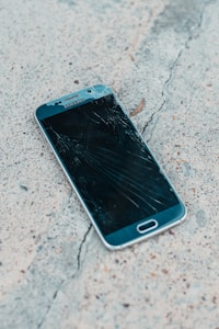A smartphone with a cracked screen lies on a rough concrete surface. The cracks spread across most of the display, indicating significant damage.