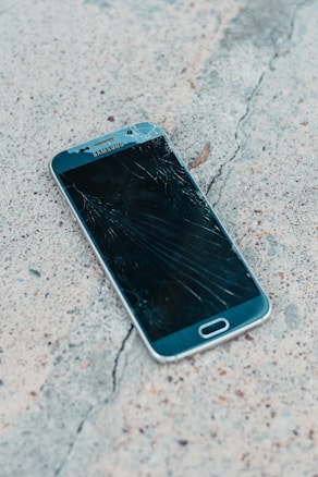 A smartphone with a cracked screen lies on a rough concrete surface. The cracks spread across most of the display, indicating significant damage.