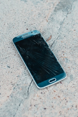 A smartphone with a cracked screen lies on a rough concrete surface. The cracks spread across most of the display, indicating significant damage.