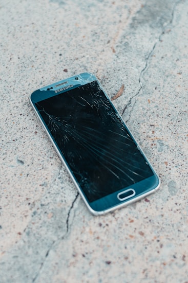 A smartphone with a cracked screen lies on a rough concrete surface. The cracks spread across most of the display, indicating significant damage.