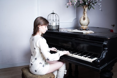 A person with long hair is playing a grand piano, adorned with a decorative birdcage and a vase of flowers. The setting includes a simple interior with a light-colored wall and wooden floor. The person is wearing a white sweater with star patterns, sitting on a cushioned bench.