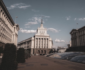 A large, imposing government building stands prominently in an open plaza, flanked by two other buildings with classical architectural elements. The sky is partly cloudy and the scene is devoid of people, giving a sense of calm and emptiness. There are manicured bushes and a glass-covered subterranean feature in the foreground.
