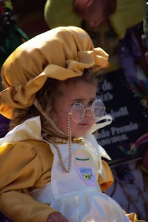 A young child is wearing a yellow bonnet and a white apron with a honey pot design. The child is also wearing round glasses and a bead necklace, looking off to the side in a setting that appears festive.