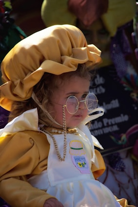 A young child is wearing a yellow bonnet and a white apron with a honey pot design. The child is also wearing round glasses and a bead necklace, looking off to the side in a setting that appears festive.
