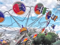 A vibrant amusement park ride features several colorful gondolas attached to an intricate, rotating framework. These circular gondolas are painted in bright reds, greens, blues, and yellows, with riders visibly seated and enjoying the experience. The ride is set against a partly cloudy sky, with trees visible in the background.