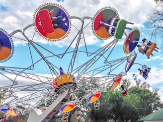 A vibrant amusement park ride features several colorful gondolas attached to an intricate, rotating framework. These circular gondolas are painted in bright reds, greens, blues, and yellows, with riders visibly seated and enjoying the experience. The ride is set against a partly cloudy sky, with trees visible in the background.