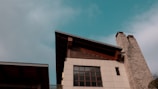 A modern building featuring a slanted roof and a tall stone chimney against a vibrant blue sky. The facade has multiple large rectangular windows and wooden accents, adding a rustic touch to the contemporary design.