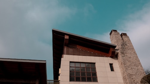 A modern building featuring a slanted roof and a tall stone chimney against a vibrant blue sky. The facade has multiple large rectangular windows and wooden accents, adding a rustic touch to the contemporary design.