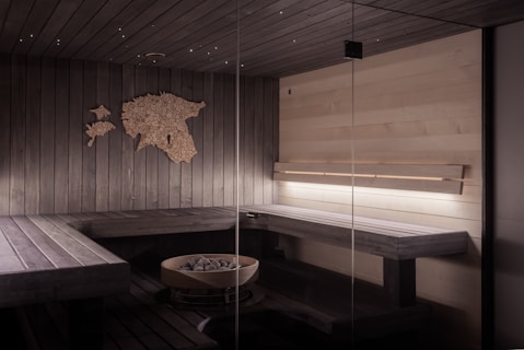 A modern sauna interior with dark wooden benches and walls. A decorative wooden map adds an artistic touch to the wall. Soft, ambient lighting illuminates the space, creating a warm and inviting atmosphere. A circular metal container filled with sauna stones is positioned on the floor.