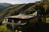 A heavily damaged and abandoned car with shattered windows is situated in a mountainous area. The surrounding landscape features green hills, tall trees, and rugged terrain under a clear blue sky.