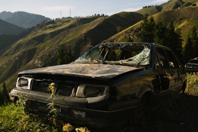 A heavily damaged and abandoned car with shattered windows is situated in a mountainous area. The surrounding landscape features green hills, tall trees, and rugged terrain under a clear blue sky.