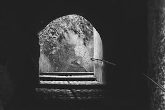 A dimly lit stone tunnel leads outward to a clearing bathed in natural light. The tunnel walls are rough and uneven, curving over a short set of stone steps. Metal handrails line the steps, guiding the path. The outdoor area features a textured stone wall with patches of moss and scattered, overhanging branches.
