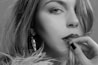 A grayscale close-up portrait of a woman with long hair. She is looking directly at the camera with a subtle, pensive expression. Her hand, with dark-colored nails, is gently touching her lips. Large, reflective earrings and textured fur or fabric are visible.