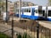 A blue and white tram marked with the number 14 and destination FlevoPark is operating on a street that is under construction. There are piles of dirt and construction equipment on the road. Several workers in orange vests are visible in the background. The foreground features a black metal fence and some green plants, and there are posters on a cylindrical structure. The background consists of traditional European buildings with multiple stories and gabled roofs.