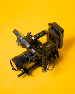 A professional camera rig setup is placed against a bright yellow background, showcasing detailed components like camera lenses, mounting brackets, and coiled cables.