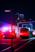 A night scene featuring an ambulance with flashing lights and a city bus on a city street. The background is urban, with tall illuminated buildings visible. The vibrant red and blue emergency lights create a striking contrast against the dark surroundings.
