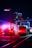A night scene featuring an ambulance with flashing lights and a city bus on a city street. The background is urban, with tall illuminated buildings visible. The vibrant red and blue emergency lights create a striking contrast against the dark surroundings.