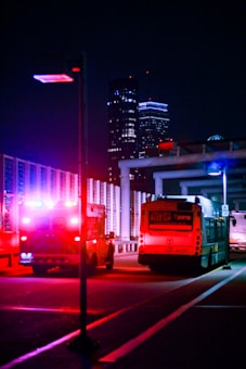 A night scene featuring an ambulance with flashing lights and a city bus on a city street. The background is urban, with tall illuminated buildings visible. The vibrant red and blue emergency lights create a striking contrast against the dark surroundings.