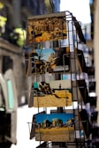 A display rack holds several colorful postcards, showcasing various scenes. The top postcard features a city skyline with historic architecture. Below it, a card displays a beach scene with people relaxing on the sand. The lower postcards also represent beachside and urban views, conveying a tourist-friendly vibe.