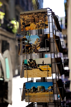 A display rack holds several colorful postcards, showcasing various scenes. The top postcard features a city skyline with historic architecture. Below it, a card displays a beach scene with people relaxing on the sand. The lower postcards also represent beachside and urban views, conveying a tourist-friendly vibe.
