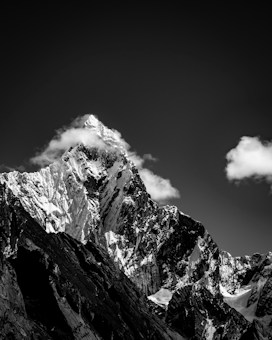 A dramatic black and white image of a sharp, snow-covered mountain peak surrounded by a few clouds under an expansive dark sky. The rugged texture of the mountain is highlighted by contrasting shadows and light.