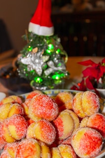 A festive setup featuring a plate of sugar-coated pastries with yellow and red colors. In the background, a small decorated Christmas tree with a red Santa hat is adorned with green lights and silver decorations. Nearby, hints of poinsettia flowers add to the holiday theme.