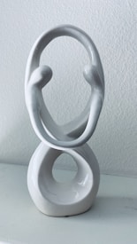 A white ceramic sculpture with an artistic, abstract design featuring two intertwined oval shapes. The top oval has two stylized human figures facing each other while the bottom forms a solid base with a hollow center. The background is a plain, light-colored wall.