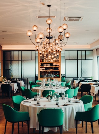 A sophisticated dining room with elegantly set tables and green upholstered chairs arranged in neat rows. A large, intricate chandelier with multiple round bulbs hangs from the ceiling, providing warm lighting. The back wall features a shelf stocked with bottles and glassware, framed by a modern, geometric design.
