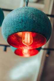 A close-up view of a textured metal pendant light with a teal exterior and bright red interior. A filament bulb hangs inside, casting a warm glow. The background is softly blurred, providing a sense of depth and focus on the light fixture.