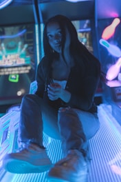 A person is sitting on a glowing, neon-lit surface in an arcade setting. The surroundings are filled with vibrant lights and arcade machines. Their casual attire includes a dark top and jeans, and the atmosphere is bathed in blue and purple hues.